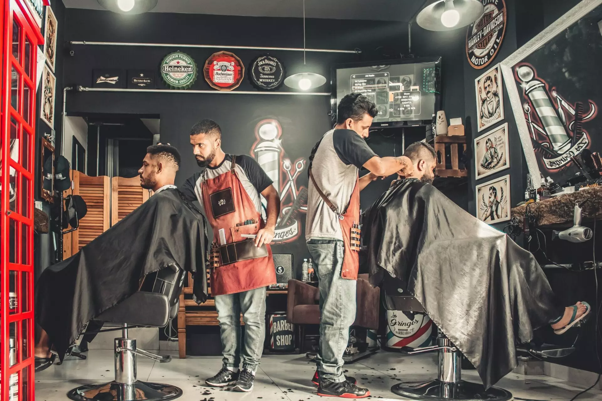 Two barbers cut hair for two seated clients in a barber shop with black walls and various posters. Both barbers wear aprons, and the clients are covered with capes. The shop is decorated with vintage barber items and lights.