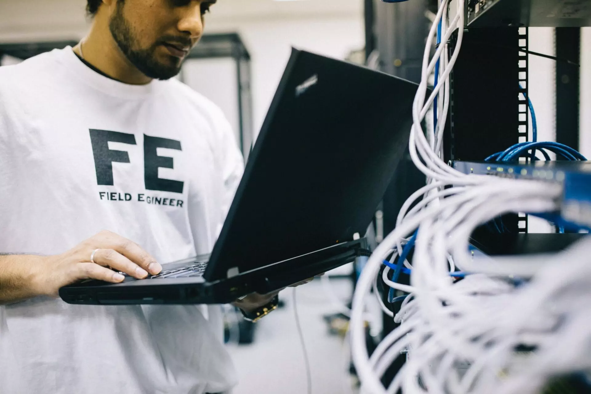 A person in a white Field Engineer t-shirt is working on a laptop. They are standing next to a server rack with various cables plugged in. The setting appears to be a data center or IT room.