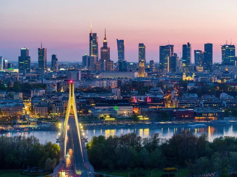 Panorama miasta o zmierzchu przedstawia nowoczesne drapacze chmur i starsze budynki. Most przecina rzekę na pierwszym planie, a światła miasta odbijają się na wodzie. W pobliżu rzeki widoczne są drzewa i drogi.
